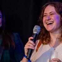 Girl is laughing into microphone while other girl smiles at er